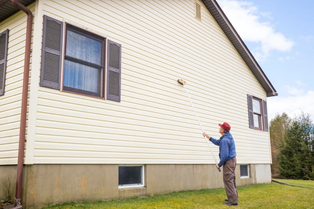 older homeowner cleans vinyl siding of home with extended brush